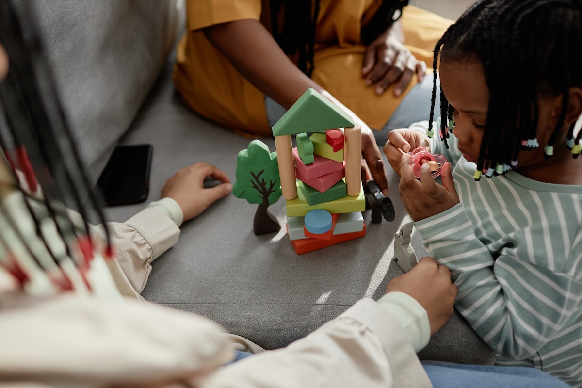Photo of a child playing with a toy, with adult childcarers assisting.