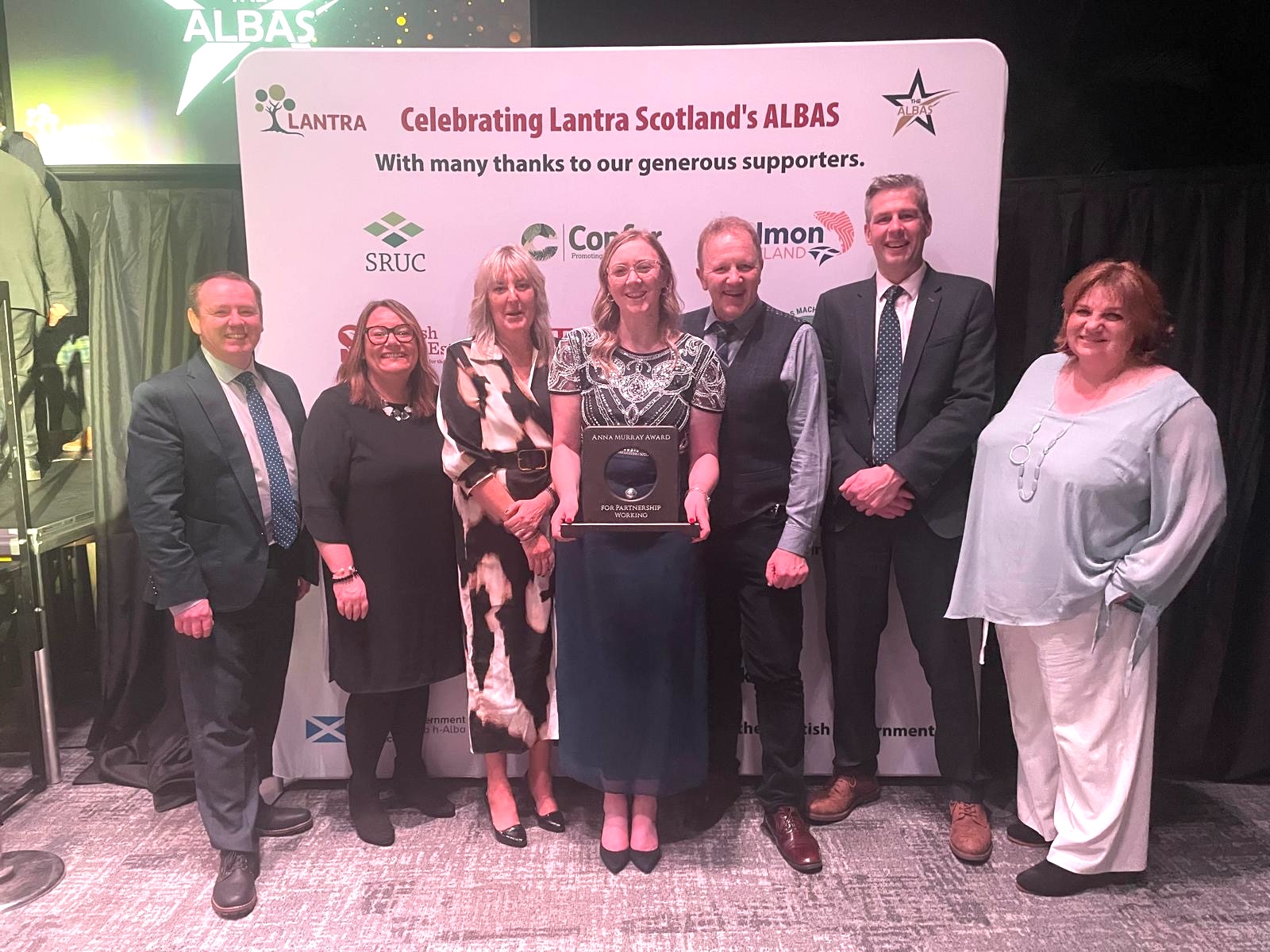 A group of seven people posing at the Lantra Scotland ALBAS awards event. They are standing in front of a backdrop featuring the Lantra logo and the words 'Celebrating Lantra Scotland’s ALBAS' along with sponsor logos.