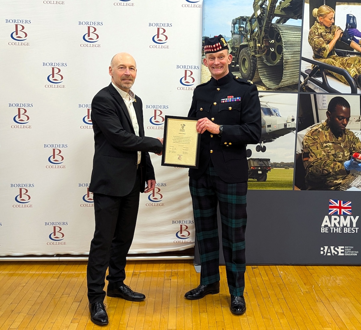 Two men shake hands while holding a certificate in front of a Borders College backdrop, with British Army promotional images in the background.