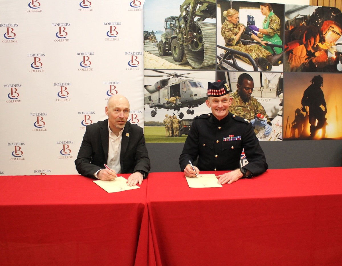 Two men sitting at a desk signing an agreement with Army images in the background