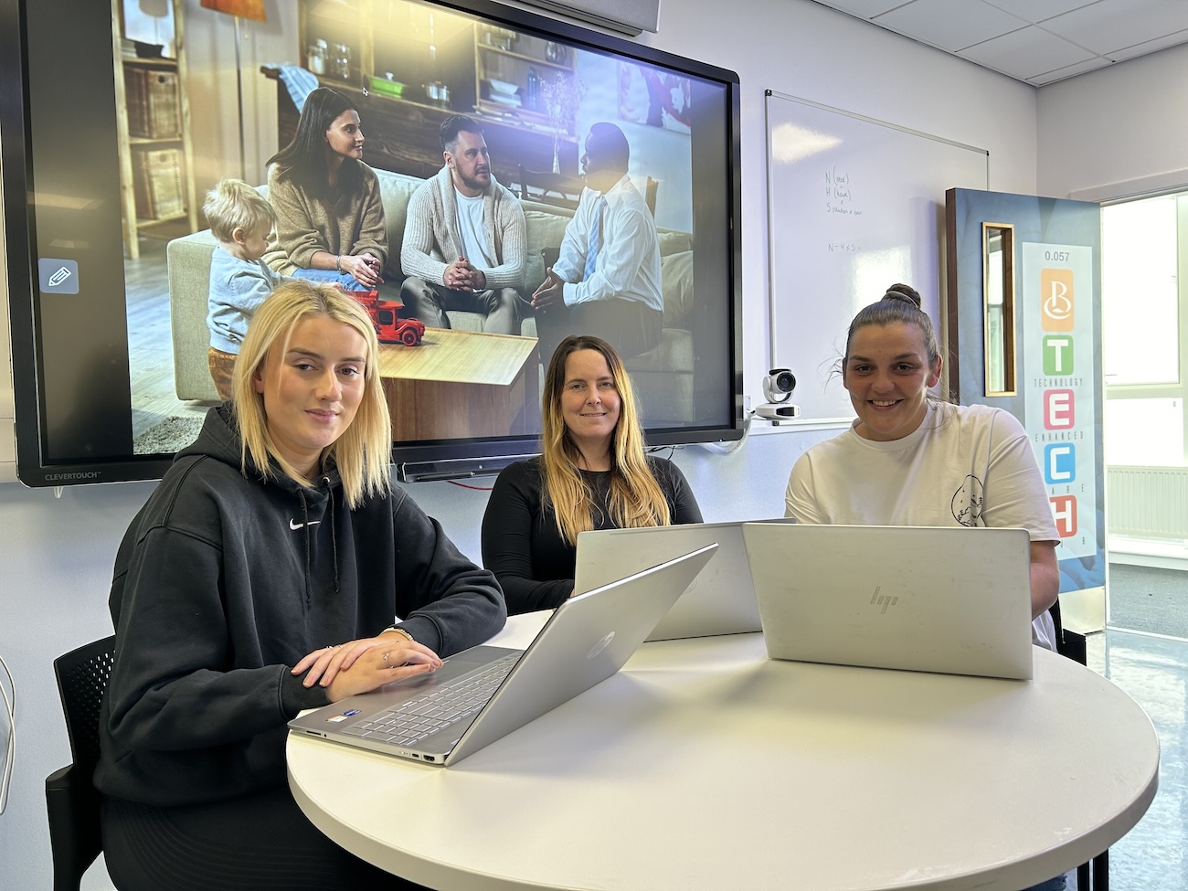 Three students sitting at a desk with laptops with a large digital display in the background.