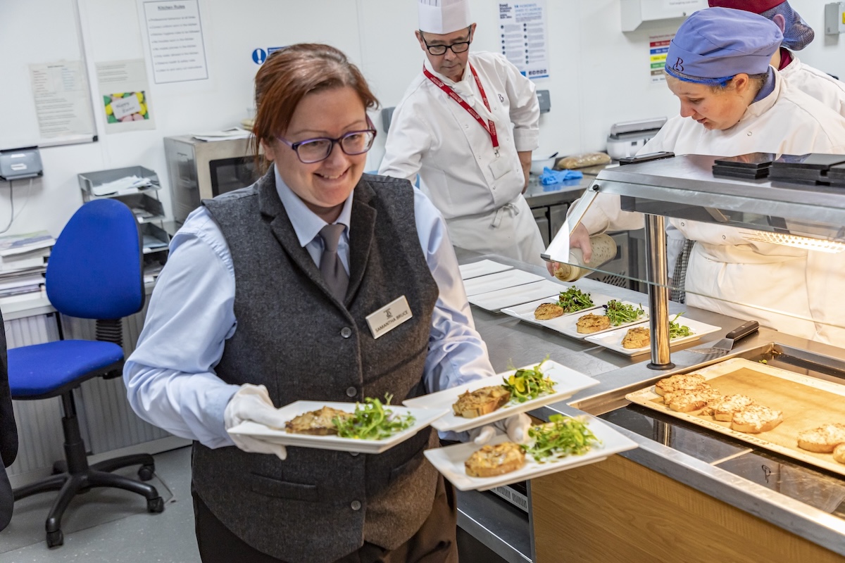 Photo of a female catering student in a kitchen setting with plates of food in her arms